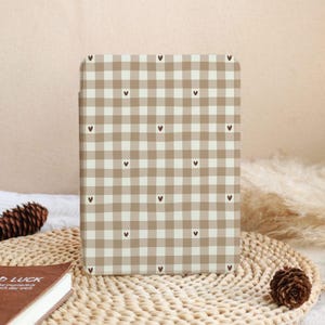 May include: A rectangular case with a tan and white checkered pattern, adorned with small brown hearts. The case is standing upright on a woven surface, with a pinecone and a book visible in the background. The case appears to be for a tablet or e-reader.