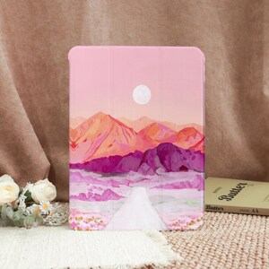 Puede incluir: Funda rosa para tableta con una pintura de paisaje de montañas en tonos naranja, morado y rosa bajo una luna blanca. Un camino blanco conduce a las montañas. La funda es un accesorio protector para tabletas.