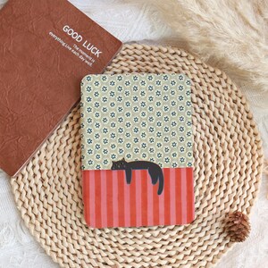 Puede incluir: Un cuaderno rectangular con un estampado floral y una secci&oacute;n inferior a rayas rojas con una ilustraci&oacute;n de gato negro. Un cuaderno marr&oacute;n con el texto "GOOD LUCK" es visible. Los art&iacute;culos est&aacute;n sobre una estera tejida.