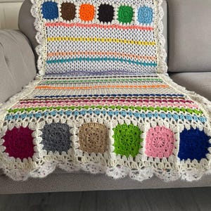 Könnte beinhalten: Handgefertigte Häkeldecke mit einer weißen Basis und verschiedenen quadratischen und gestreiften Mustern. Die Decke hat Quadrate in Blau, Braun, Orange, Schwarz, Grün und Rosa, mit einem gewellten Rand. Die Decke liegt auf einem Sofa.