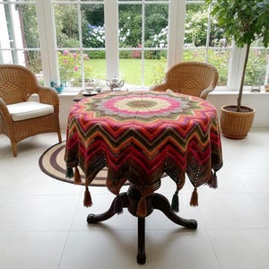 Könnte beinhalten: Ein runder Tisch mit einer farbenfrohen, gemusterten Häkeltischdecke. Die Tischdecke hat ein Zickzackmuster in Rot-, Orange-, Grün- und Brauntönen mit Quasten an den Kanten. Zwei Korbstühle und eine Topfpflanze sind im Hintergrund zu sehen.
