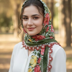 May include: A floral headscarf with a green border and a white background adorned with red and yellow flowers. The scarf has a crocheted edge in a reddish-brown color. The person is wearing a white blouse.