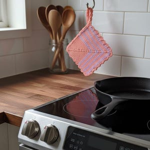 May include: A handmade, knitted pot holder in shades of peach and lavender hangs from a hook in a kitchen. The pot holder has a scalloped edge and a striped pattern. A black cast iron skillet sits on a stovetop. Wooden spoons are in a glass jar.