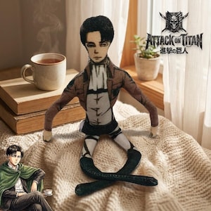 Op de afbeelding: Een pluche pop van een personage uit "Attack on Titan" met een bruine jas, wit overhemd en zwarte broek. De pop zit op een beige deken, met een kopje thee en boeken op de achtergrond. Het "Attack on Titan"-logo is zichtbaar.