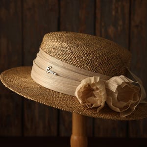 Puede incluir: Un sombrero de paja color canela con copa plana y ala ancha. Una cinta beige envuelve la base de la copa, con dos flores de tela plisada en el lateral y un pequeño dije de metal plateado con la palabra Grace en letra cursiva.