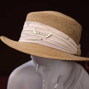 May include: A beige straw hat with a wide brim and a cream-coloured band. The band features a silver-coloured pin with the name "Eleanor". The hat is displayed on a white mannequin head against a dark background.