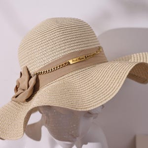 Op de afbeelding: Een beige strooien zonnehoed met een brede rand en een decoratieve band. De band heeft een gouden ketting en een klein rechthoekig label met de tekst "Best Mom". Een stoffen bloem siert de zijkant van de hoed.