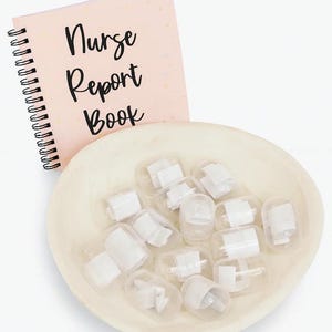 Puede incluir: Un cuaderno rosa con espiral con las palabras "Nurse Report Book" en escritura negra. Un cuenco poco profundo, blanquecino, contiene varios recipientes de pl&aacute;stico transparente peque&ntilde;os con contenido blanco. Los art&iacute;culos est&aacute;n dispuestos sobre una superficie blanca.