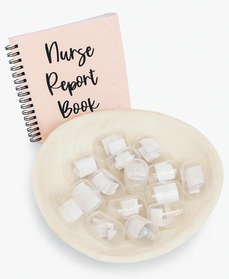Puede incluir: Un cuaderno rosa con espiral con las palabras "Nurse Report Book" en escritura negra. Un cuenco poco profundo, blanquecino, contiene varios recipientes de pl&aacute;stico transparente peque&ntilde;os con contenido blanco. Los art&iacute;culos est&aacute;n dispuestos sobre una superficie blanca.