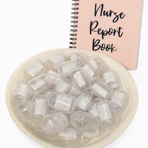 May include: A light pink spiral-bound notebook with the words "Nurse Report Book" in black script sits next to a wooden bowl filled with clear, capsule-shaped containers. The containers have white paper inserts.