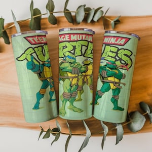 May include: Three green stainless steel tumblers with Teenage Mutant Ninja Turtles graphics. Each tumbler has a different character illustration and the words "Teenage Mutant Ninja Turtles" in a bold, green font. The tumblers are cylindrical and have a silver rim.