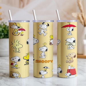 May include: Three cylindrical yellow tumblers with clear lids and white straws. The tumblers feature a repeating pattern of Snoopy and Woodstock in various poses, including holding umbrellas, sitting on a doghouse, and the text SNOOPY in orange letters.