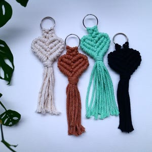 Puede incluir: Cuatro llaveros de macramé en crema, marrón, verde azulado y negro. Cada llavero presenta un diseño de nudo en forma de corazón con una borla. Los llaveros están dispuestos sobre una superficie blanca, con una planta verde en la esquina superior izquierda.