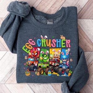 May include: Dark gray sweatshirt with the words "EGG CRUSHER" in colorful letters. The graphic features monster trucks with superhero characters and Easter eggs. The sweatshirt is folded on a light wood-grain surface.