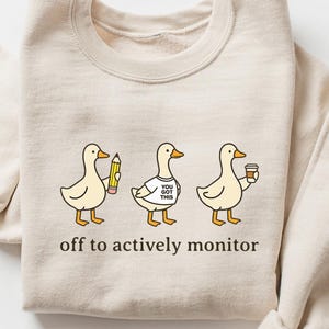 May include: A beige sweatshirt with a crew neck and rolled-up sleeves. The front features a cartoon illustration of three ducks, each holding an item. The text "off to actively monitor" is printed below the ducks.