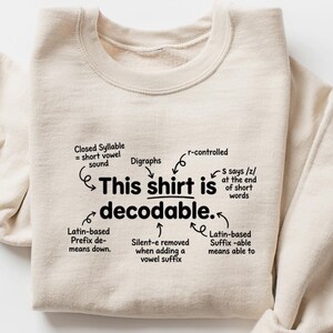 May include: A cream-colored crewneck sweatshirt featuring black text that reads, "This shirt is decodable." The design includes various linguistic annotations with arrows pointing to specific words, explaining concepts like closed syllables, digraphs, r-controlled vowels, and Latin-based prefixes and suffixes.