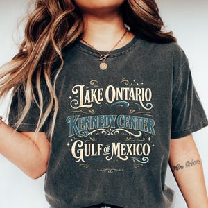 May include: A charcoal grey short-sleeve t-shirt featuring a graphic print with the text Lake Ontario, Kennedy Center, and Gulf of Mexico in a decorative font style, accented with small stars and flourishes in cream and blue colors.