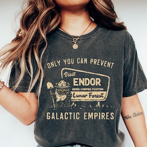 May include: Dark grey t-shirt with the text "ONLY YOU CAN PREVENT Visit ENDOR Hiking-Camping-Feasting Lunar Forest GALACTIC EMPIRES." The shirt has a vintage-style graphic of a ranger. The person is wearing blue jeans and white shoes.