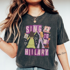 May include: A charcoal gray short-sleeve t-shirt featuring a graphic print with the text "SING TO ME HILARY" in purple blocks. The design includes two figures, a red scooter, musical notes, and small decorative icons in various colors.
