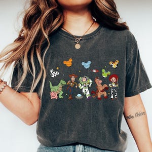 Pu&ograve; includere: T-shirt grigio scuro con una grafica colorata dei personaggi di Toy Story, tra cui Woody e Buzz Lightyear. Il design presenta anche altri personaggi del film. La maglietta &egrave; abbinata a jeans blu e scarpe da ginnastica bianche.