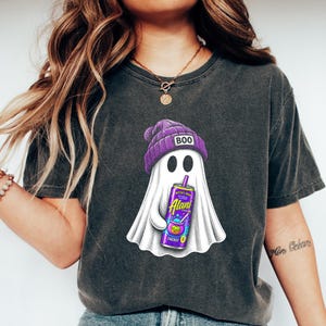 May include: A charcoal gray t-shirt featuring a graphic of a white ghost wearing a purple beanie with the text BOO on it. The ghost is holding a purple Alani energy drink can labeled Witch's Brew. The shirt is paired with blue denim jeans.