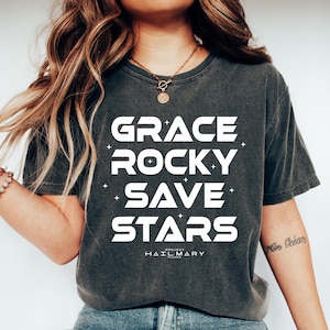 May include: Dark gray t-shirt with the words "GRACE ROCKY SAVE STARS" in large white letters. Below the words is the text "HAIL MARY" in smaller white letters. The shirt is paired with blue jeans and white sneakers.