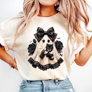 May include: A cream-colored t-shirt featuring a graphic print of a ghost wearing a large black lace bow and holding a small black kitten. The design is surrounded by black roses and lace trim, creating a detailed illustration on the front of the shirt.