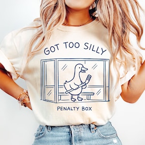 K&ouml;nnte beinhalten: Cremefarbenes T-Shirt mit dem Text "GOT TOO SILLY" &uuml;ber einer blauen Grafik einer Ente in einer Strafbox, die einen Hockeyschl&auml;ger h&auml;lt. Die Worte "PENALTY BOX" stehen darunter. Das Shirt wird mit hellblauen Jeans getragen.