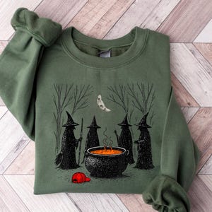 May include: An olive green crewneck sweatshirt featuring a graphic print of four black-robed figures in pointed hats standing around a bubbling orange cauldron in a forest under a crescent moon, with a small red baseball cap on the ground.