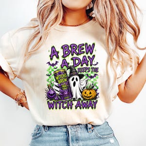 Peut inclure: Un t-shirt couleur crème avec une impression graphique portant le texte A BREW A DAY KEEPS THE WITCH AWAY. Le motif comprend un fantôme portant un chapeau de sorcière, une canette étiquetée WITCH'S BREW, une potion violette, une citrouille et des chauves-souris.