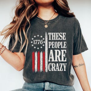 May include: Dark gray t-shirt featuring a distressed American flag design with the text "1776" and "These People Are Crazy" in white. The shirt is worn with blue jeans and white sneakers.