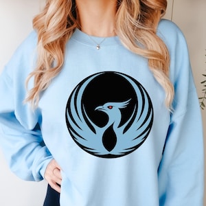 May include: A light blue sweatshirt featuring a black and light blue phoenix design. The phoenix is encircled by a black circle. The bird has red eyes and is centered on the front of the sweatshirt.
