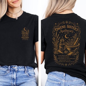 May include: A black t-shirt featuring a gold graphic of a mortar and pestle, herbs, and a crescent moon. The front chest displays a small version of the design, while the back features a larger version with the text Owens Sister's Apothecary.