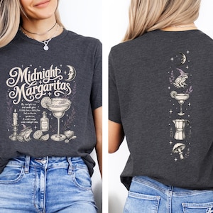 May include: A dark gray t-shirt featuring a front graphic with the text Midnight Margaritas, a margarita glass, a candle, a frog in a witch hat, and a bottle. The back graphic displays a vertical arrangement of a crescent moon, a frog, a margarita, a coffee pot, and a mushroom.