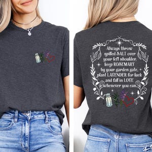 May include: A dark gray t-shirt featuring a small graphic on the front left chest of a salt shaker, rosemary sprig, lavender, and a red heart. The back displays the same icons within a floral wreath and the text: Always throw spilled SALT over your left shoulder, keep ROSEMARY by your garden gate, plant LAVENDER for luck and fall in LOVE whenever you can.