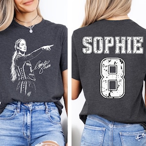 May include: A dark gray t-shirt featuring a white graphic of a basketball player pointing on the front and the name SOPHIE with the number 8 in a distressed white font on the back. The shirt is paired with light blue distressed denim jeans.