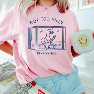 K&ouml;nnte beinhalten: Hellrosa Sweatshirt mit dem Text "GOT TOO SILLY" und einer Cartoon-Ente in einer Strafbox mit einem Hockeyschl&auml;ger. Das Sweatshirt wird mit hellblauen Shorts getragen. Eine wei&szlig;e Tasse mit einem gelben Smiley wird gehalten.