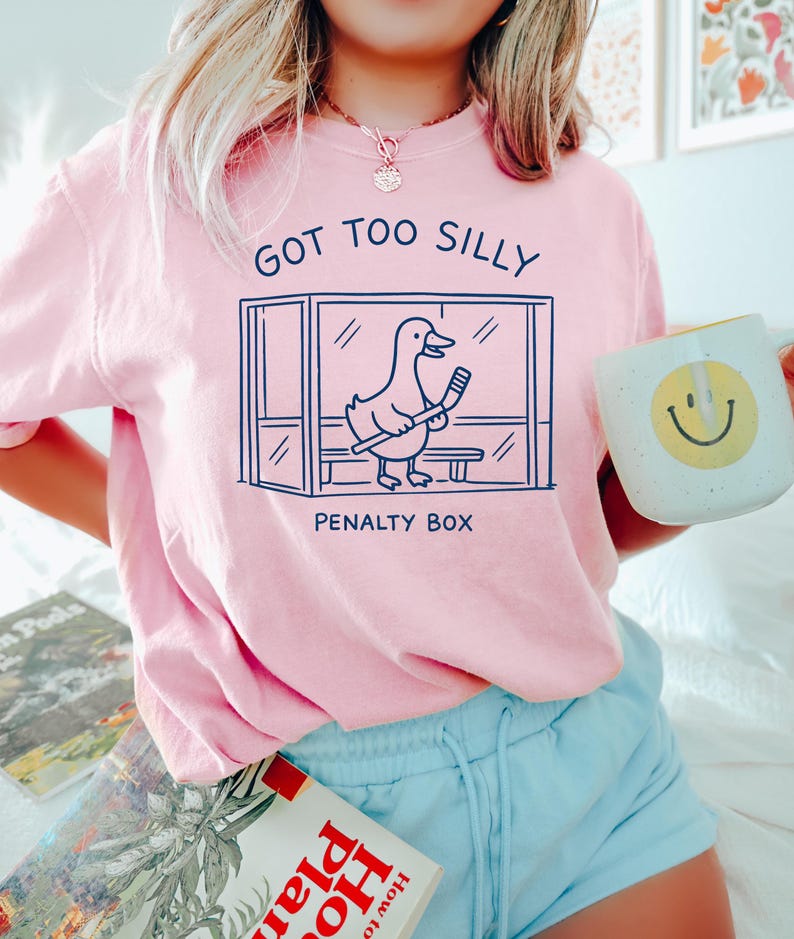 K&ouml;nnte beinhalten: Hellrosa Sweatshirt mit dem Text "GOT TOO SILLY" und einer Cartoon-Ente in einer Strafbox mit einem Hockeyschl&auml;ger. Das Sweatshirt wird mit hellblauen Shorts getragen. Eine wei&szlig;e Tasse mit einem gelben Smiley wird gehalten.