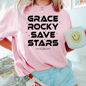 May include: A light pink sweatshirt with the words "GRACE ROCKY SAVE STARS" in bold black letters. Below the text is "PROJECT HAIL MARY". A white mug with a yellow smiley face is held in the hand.