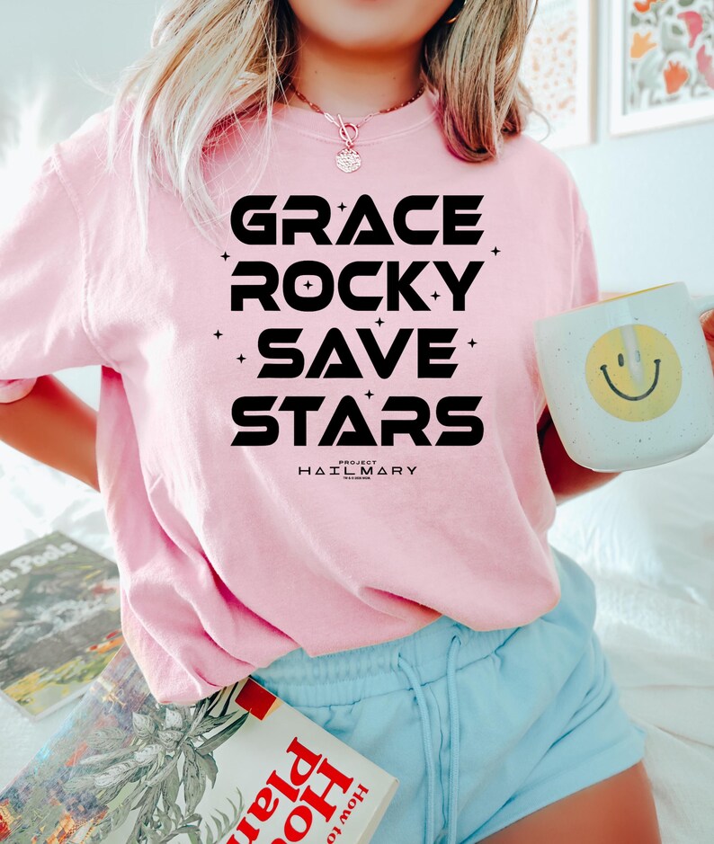 May include: A light pink sweatshirt with the words "GRACE ROCKY SAVE STARS" in bold black letters. Below the text is "PROJECT HAIL MARY". A white mug with a yellow smiley face is held in the hand.