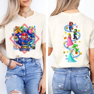 May include: Cream-colored t-shirt featuring a colorful graphic of Mario and other characters from the Super Mario Galaxy game. The back of the shirt displays additional characters. The text "THE SUPER MARIO GALAXY" is also visible.
