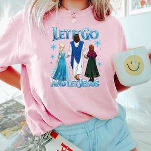 Puede incluir: Camiseta rosa claro con el texto azul y blanco "LET IT GO AND LET JESUS". El gr&aacute;fico presenta dos chicas con vestidos y una figura con una t&uacute;nica azul. Una prenda casual para el uso diario.