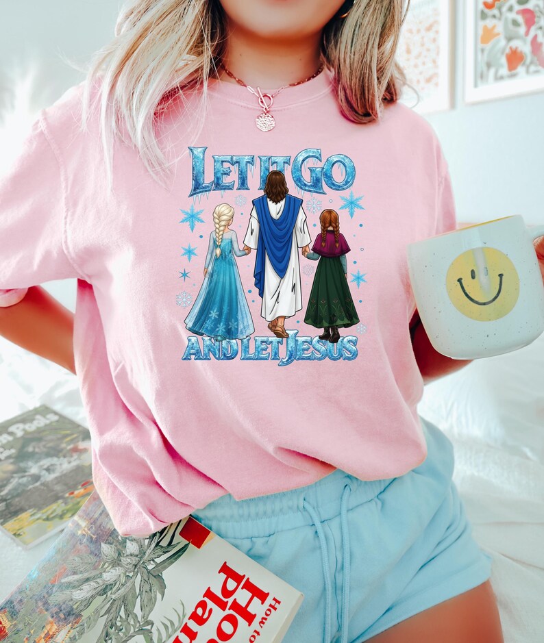 Puede incluir: Camiseta rosa claro con el texto azul y blanco "LET IT GO AND LET JESUS". El gr&aacute;fico presenta dos chicas con vestidos y una figura con una t&uacute;nica azul. Una prenda casual para el uso diario.