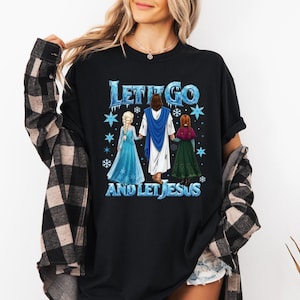 Puede incluir: Camiseta negra con el texto "LET IT GO AND LET JESUS" en azul y blanco. El gr&aacute;fico muestra a Jes&uacute;s de la mano de dos personajes de dibujos animados en una escena invernal.