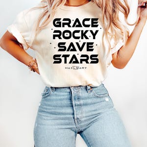 May include: Cream-colored t-shirt with the words "GRACE ROCKY SAVE STARS" in bold black letters. The text is arranged vertically with small star accents. The brand name "HAIL MARY" is printed below the text. The shirt is paired with light blue jeans.