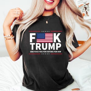 May include: Black t-shirt with the text "F**K TRUMP" in white block letters, featuring the American flag. The text "AND F**K YOU FOR VOTING FOR HIM" is below the main text in red.