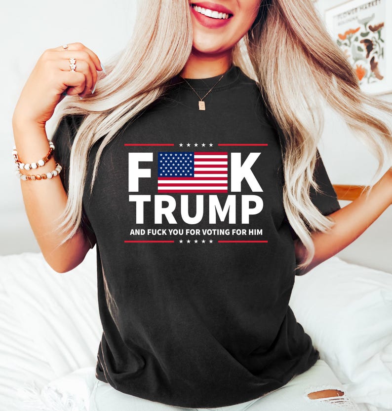 May include: Black t-shirt with the text "F**K TRUMP" in white block letters, featuring the American flag. The text "AND F**K YOU FOR VOTING FOR HIM" is below the main text in red.