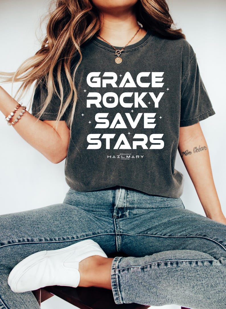 May include: Dark gray t-shirt with the words "GRACE ROCKY SAVE STARS" in large white letters. Below the words is the text "HAIL MARY" in smaller white letters. The shirt is paired with blue jeans and white sneakers.