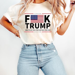 May include: Cream-colored t-shirt with the text "F**K TRUMP" in bold black letters, featuring an American flag design. The text "AND F**K YOU FOR VOTING FOR HIM" is printed below. The shirt is paired with light blue jeans.