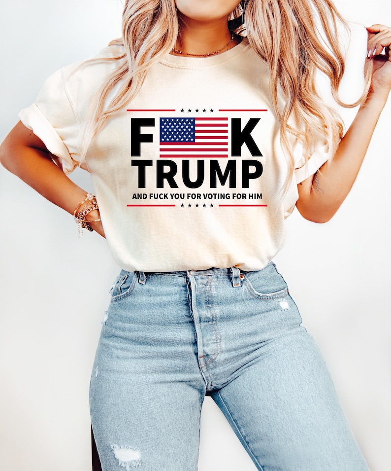 May include: Cream-colored t-shirt with the text "F**K TRUMP" in bold black letters, featuring an American flag design. The text "AND F**K YOU FOR VOTING FOR HIM" is printed below. The shirt is paired with light blue jeans.